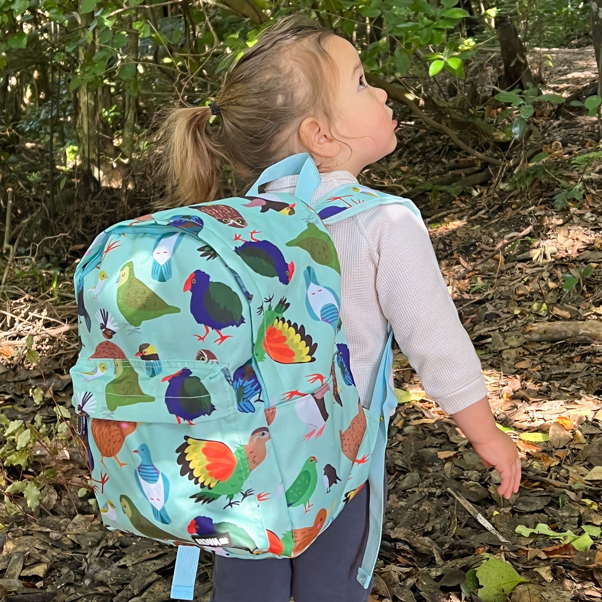 Young girl walking in the bush wearing a Minty green backpack covered in bright prints of New Zealand birds.