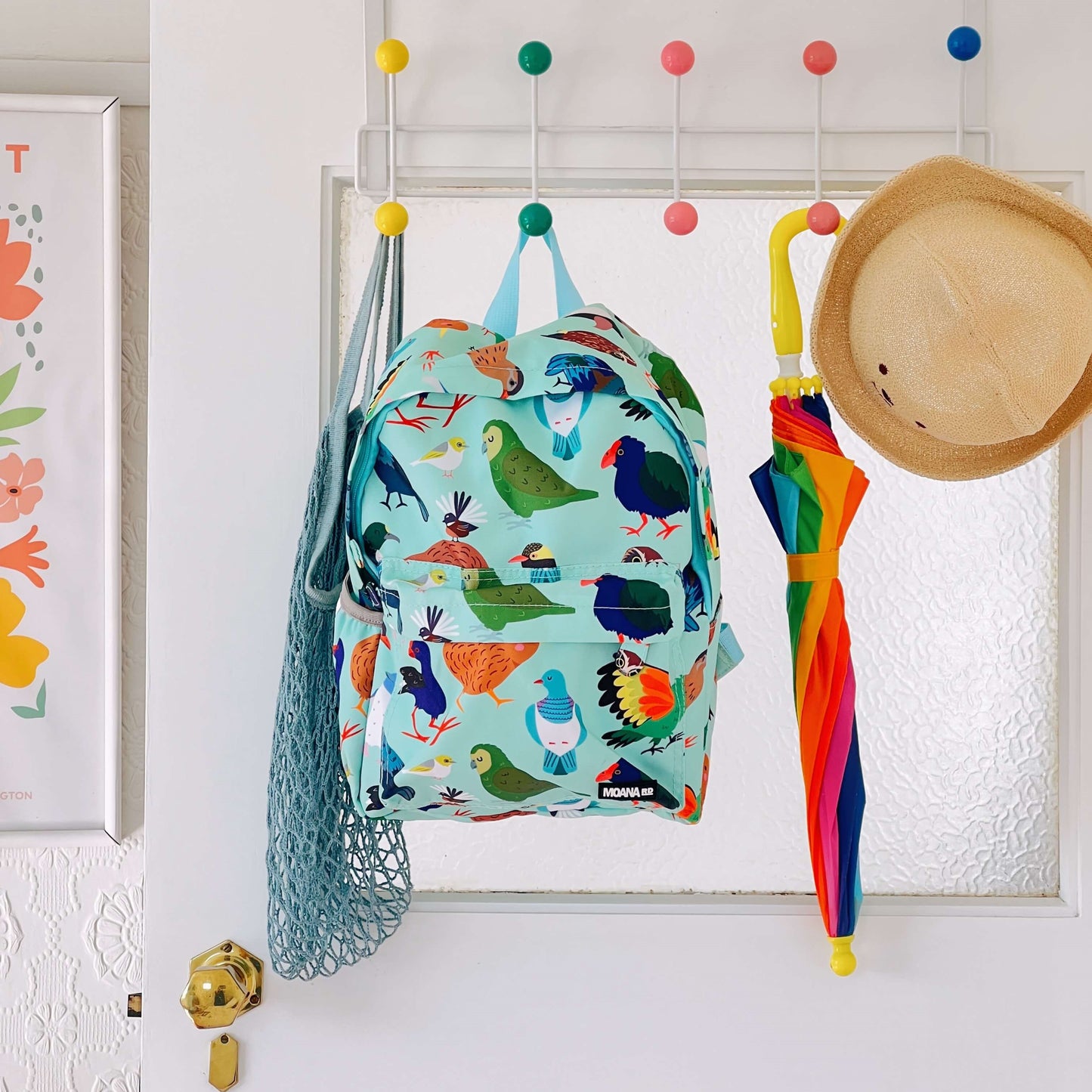 Minty green backpack covered in bright prints of New Zealand birds. The bag is hanging on a coloured hook next to a rainbow umbrella, straw hat and blue net bag.