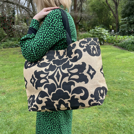 Close up of a jute tote bag with black damask style print over the shoulder of a person in a black and green dress amongst a green garden setting.