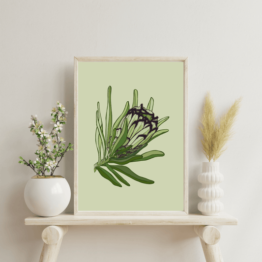 Digital art by Bridget King featuring a Green and black minky protea flower on a pale grey background. Framed in a light wooden frame sitting on a wooden side table with a white vase either side containing flowers.