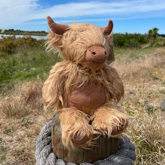 A soft toy Highland Cow sitting on a fence post wiith an estuary view behind it. The toy has a shaggy light brown fur, darker brown horns, nose and tummy. 