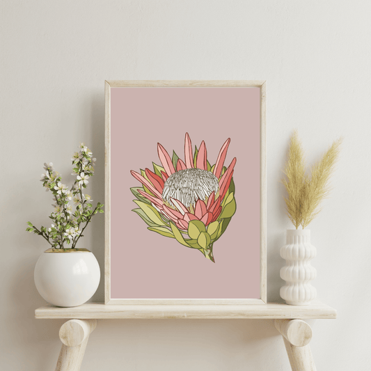 Digital print artwork by Bridget King featuring a pink King Protea flower. Framed in a light wooden frame sitting on a wooden side table with a white vase either side containing flowers.