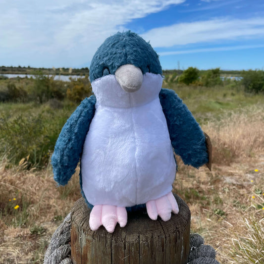 Soft toy little blue penguin with white tummy and blue head and flippers sitting on a fence post with an estaury view in the background.