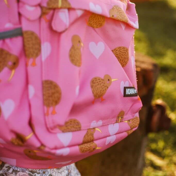 Close up of the front pocket of a pink kids backpack with lighter pink hearts and brown kiwi birds print.