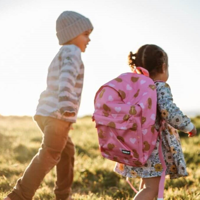 A boy walking outdoors next to a girl who is wearing a pink kids backpack with lighter pink hearts and brown kiwi birds print.