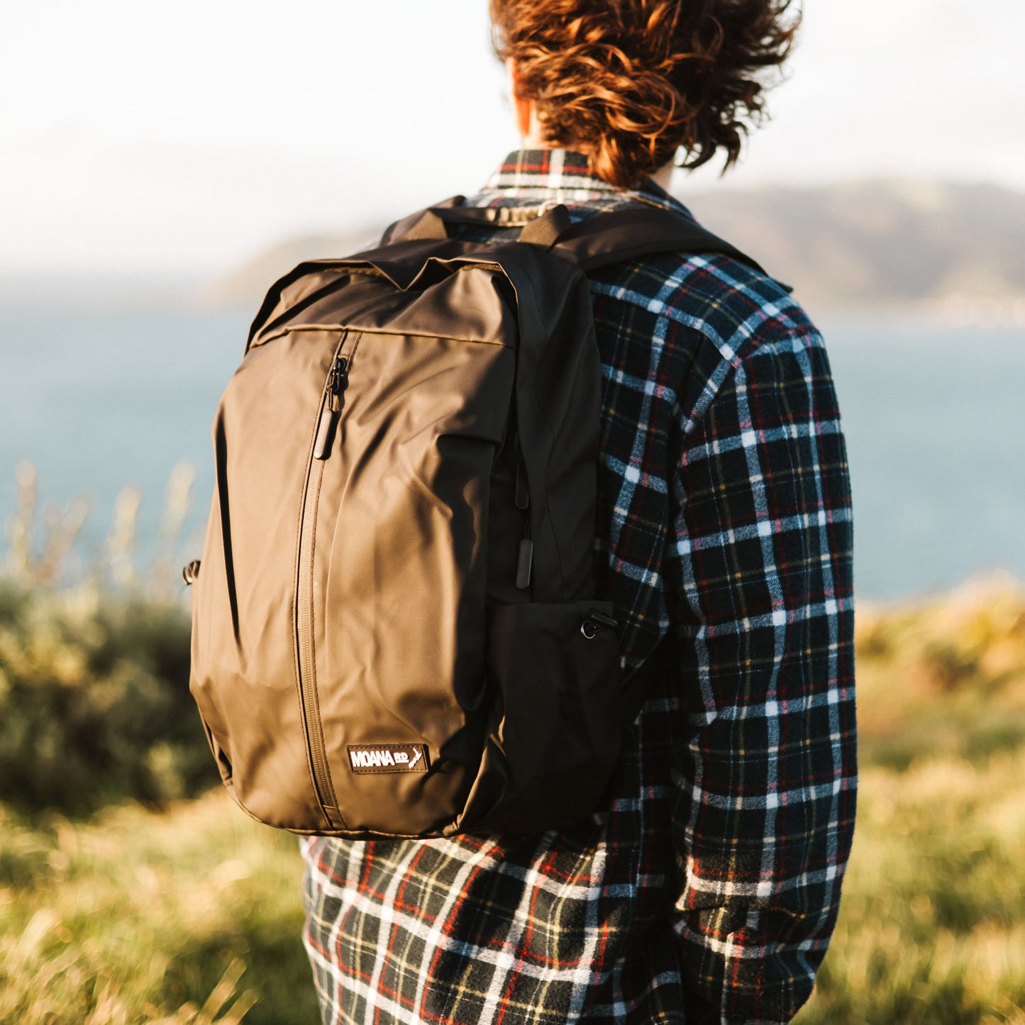 Man in plaid shirt overlooking the ocean and wearing a black Moana RD backpack.