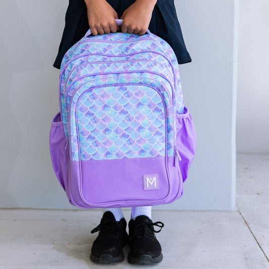 Childs hands holding top strap of a purple backpack with a blue and purple toned mermaid scale print.