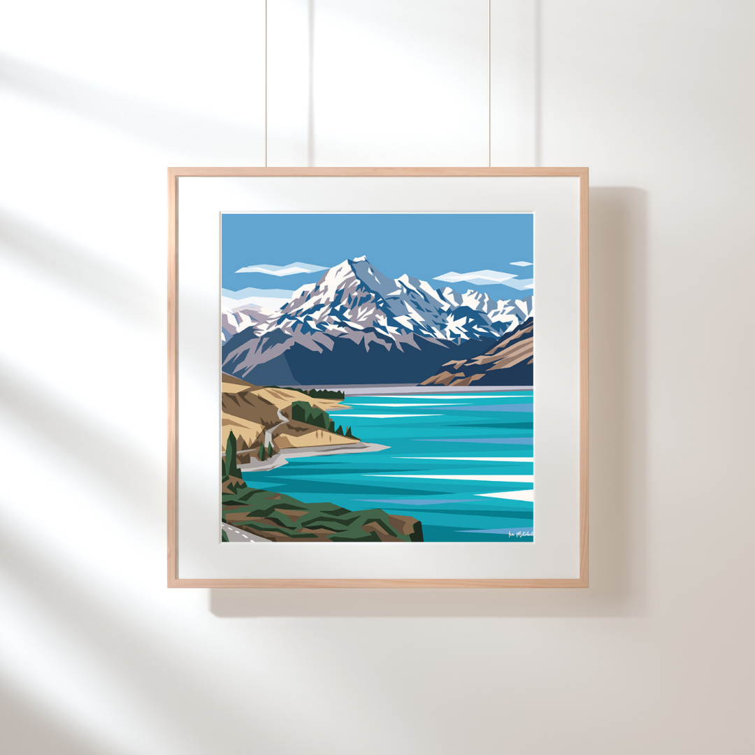 Art print by Ira Mitchell. Geometric style image featuring a snow capped Mt Cook in the background with a bright blue Lake Pukaki in the foreground. Hanging against a white wall in a light wooden frame with white border.