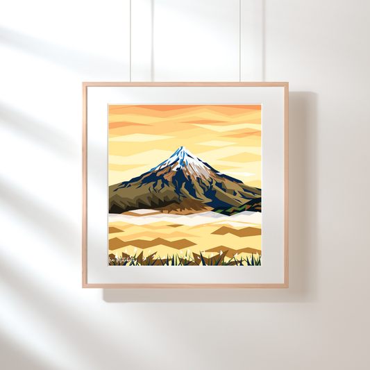 Art print by Ira Mitchell. Geometric style image featuring a snow capped Mt Taranaki at Sunrise in brown and orange hues. Hanging against a white wall in a light wooden frame with white border.