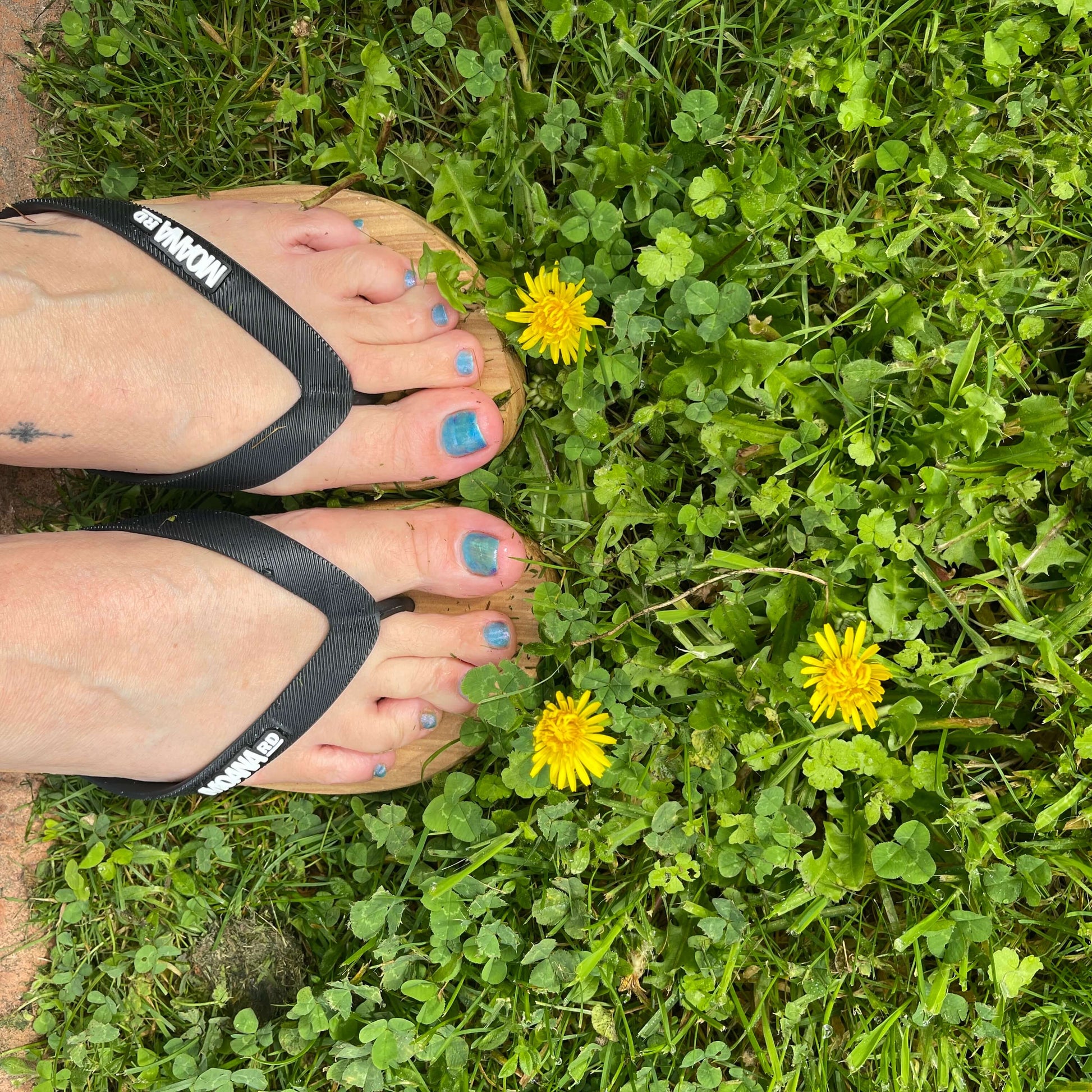 Womens feet wearing woodgrain look jandals with black straps on some grass with yellow dandelion flowers.