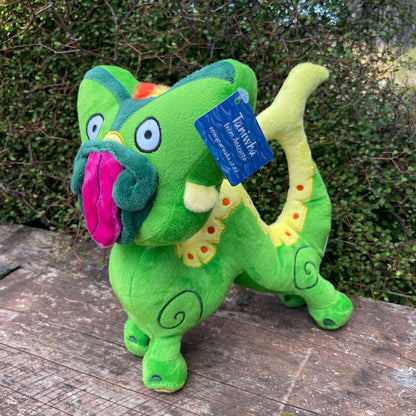 Bright green Taniwha soft toy with big pink tongue and yellow tail sitting on a wooden bench with green hedge behind it.