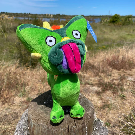 Bright green Taniwha soft toy with big pink tongue and yellow tail sitting on a fence post with an estuary in the background.