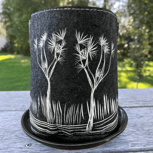 Dark grey felt plant eco plant pot with white Ti Kouka tree printed on it, sitting in a matching saucer with a blurred garden in the background.