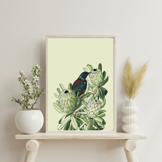 Digital art print by Bridget King featuring a Tui bird nestled amongst protea flowers. Framed in a light wooden frame sitting on a wooden side table with a white vase either side containing flowers.