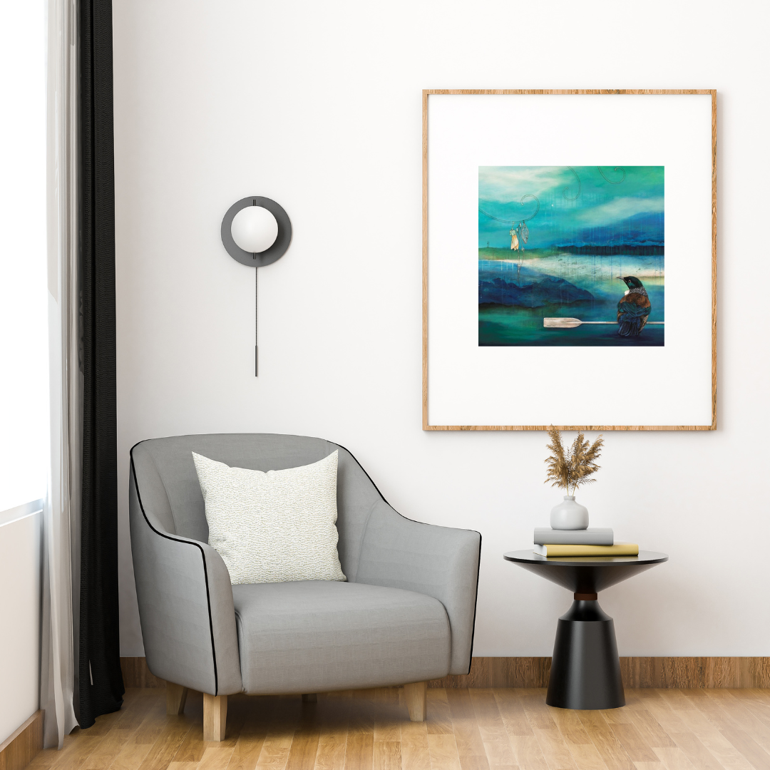 Art print by Kathryn Furniss featuring a Tui bird perched on a paddle with a blue and green background of a small enclosed bay. There is a sketch of a kowhai flower hanging from a koru o the left. The print is in a wooden frame with large white border on a white wall in a corner of a room with a grey chair and small side table.