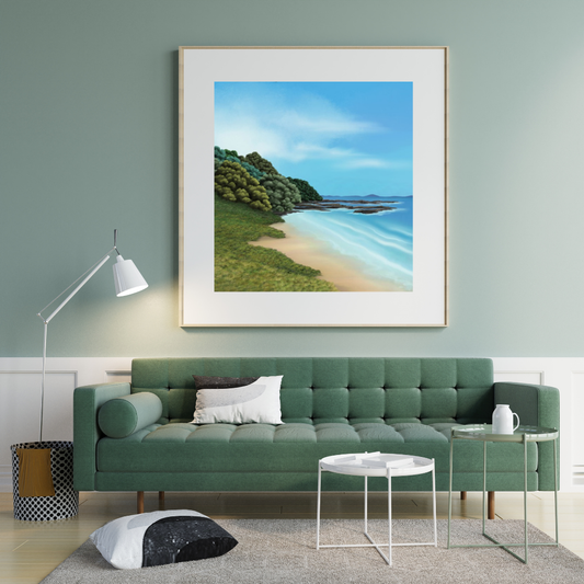 Art print of Waipapa Cable Bay by Anna Evans in a large square wooden frame with white border, hanging on light grey wall above a light gree velvet couvh with white coffee table and lamp.