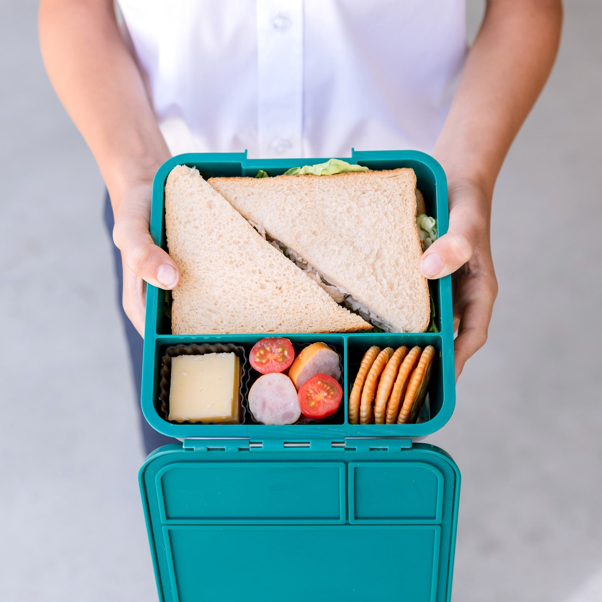 Childs hands holding a teal green bento style lunchbox with 3 compartments. These are filled with sandwiches, crackers, cheese and tomatoes.