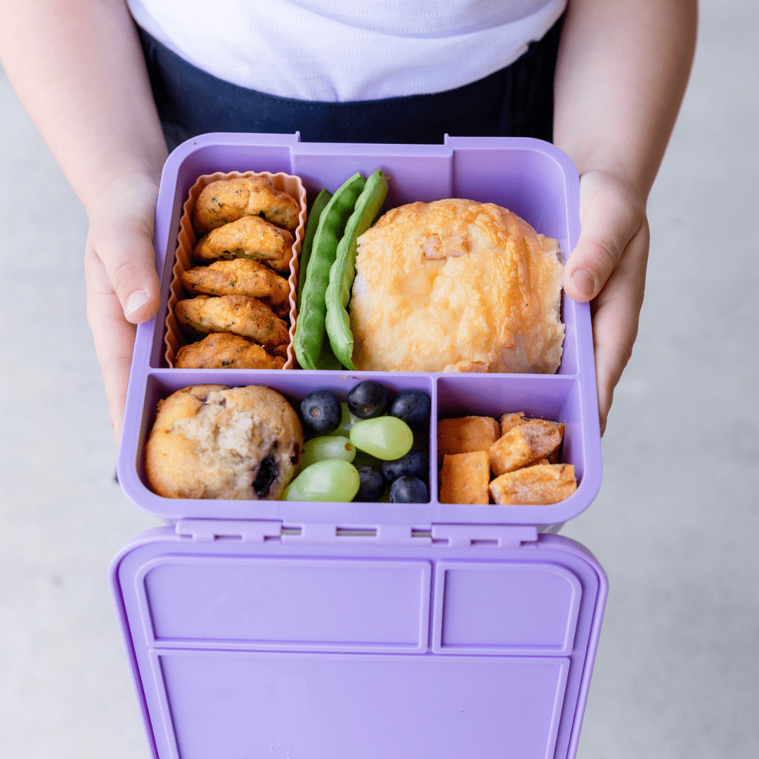 Close up of a lilac purple bento style lunchbox held by a child. Inside the lunchbox is a bun, peas, cookies, muffin and grapes.