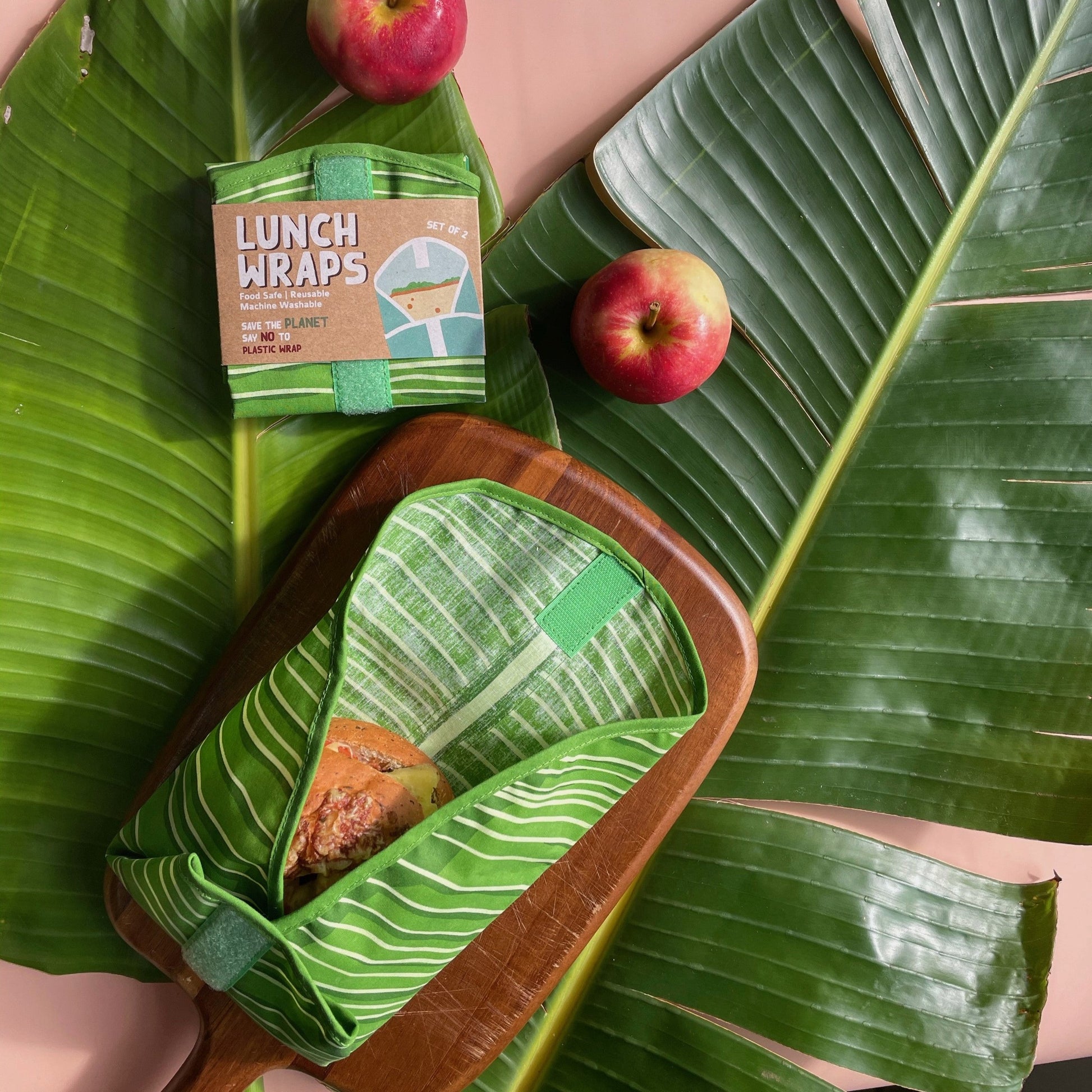 Lunch wraps in a leaf pattern containing a sandwich sitting on top of large leaves with some apples.