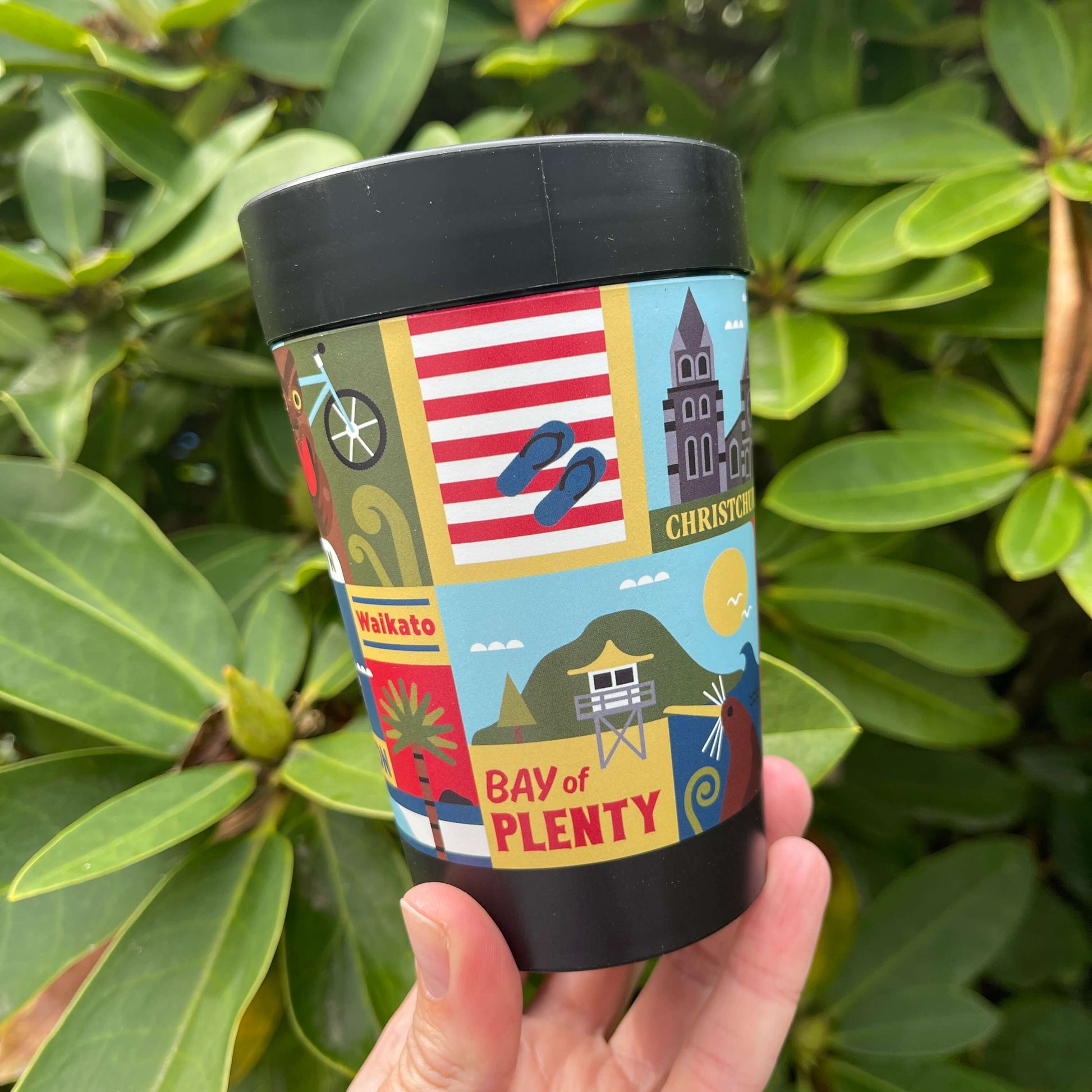 Reusable coffee cup in black with artist images of New Zealand towns wrapped around it.