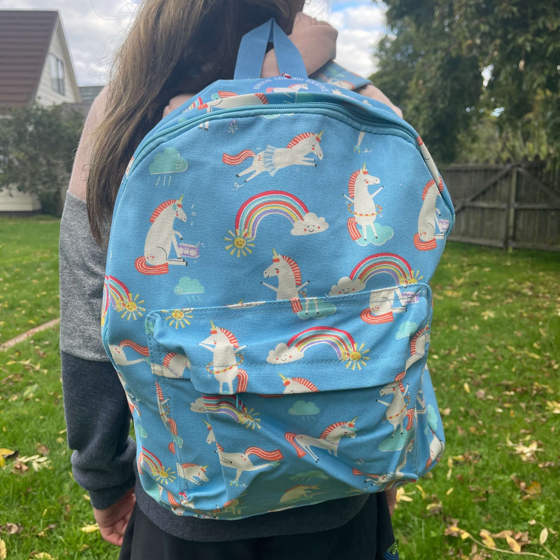 Girl wearing a sky blue backpack with unicorns and rainbows printed on it.