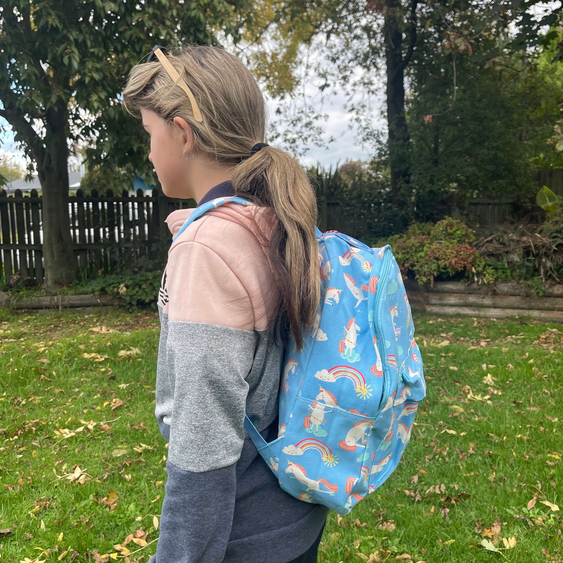 Girl wearing a sky blue backpack with unicorns and rainbows printed on it.